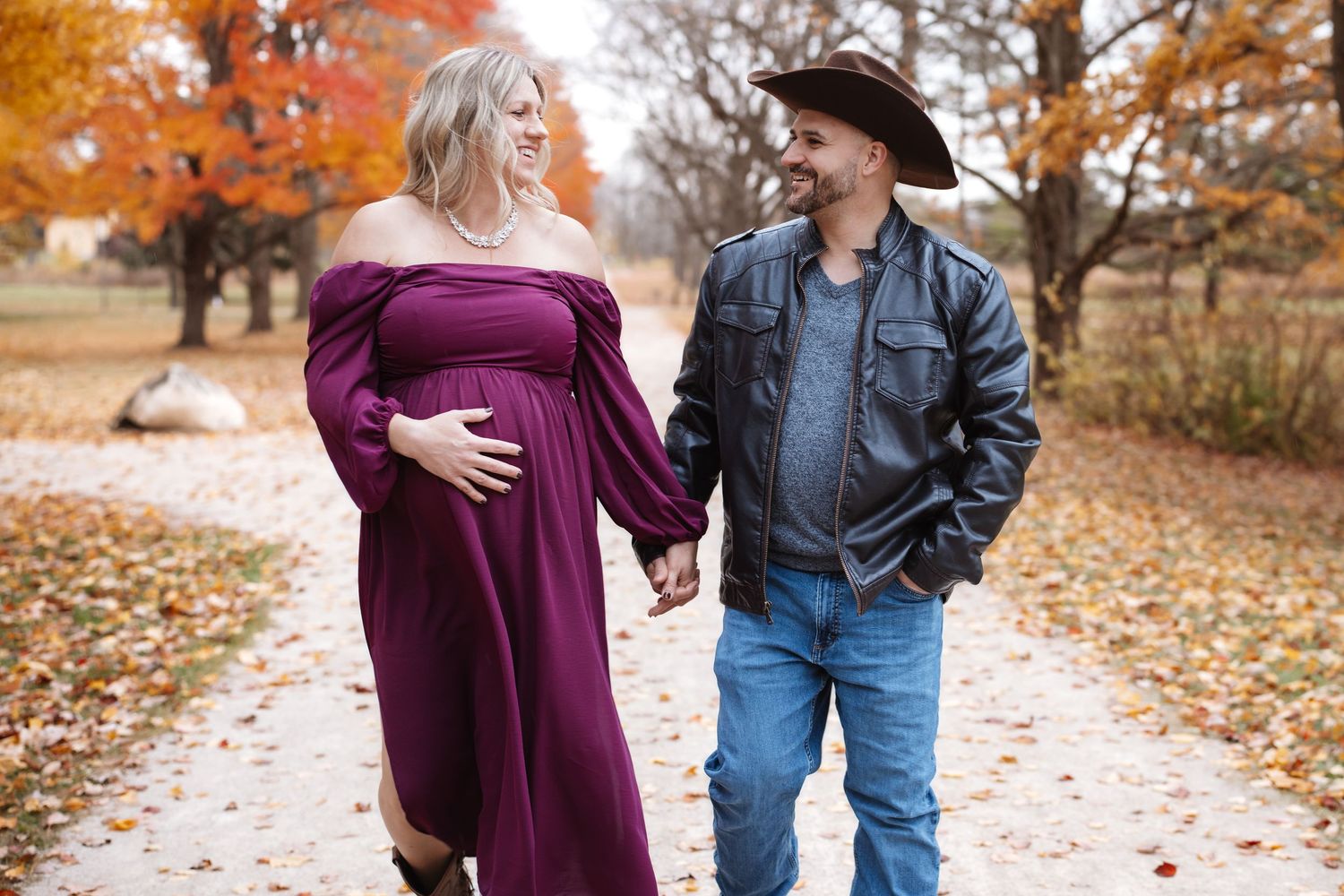 Happy expecting couple walking hand-in-hand on a leaf-strewn path with colorful autumn trees.