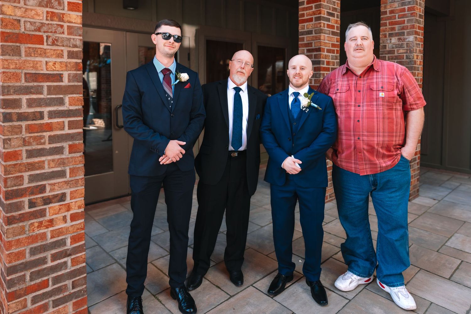Four men in suits and casual attire pose together outside a brick building at a wedding.