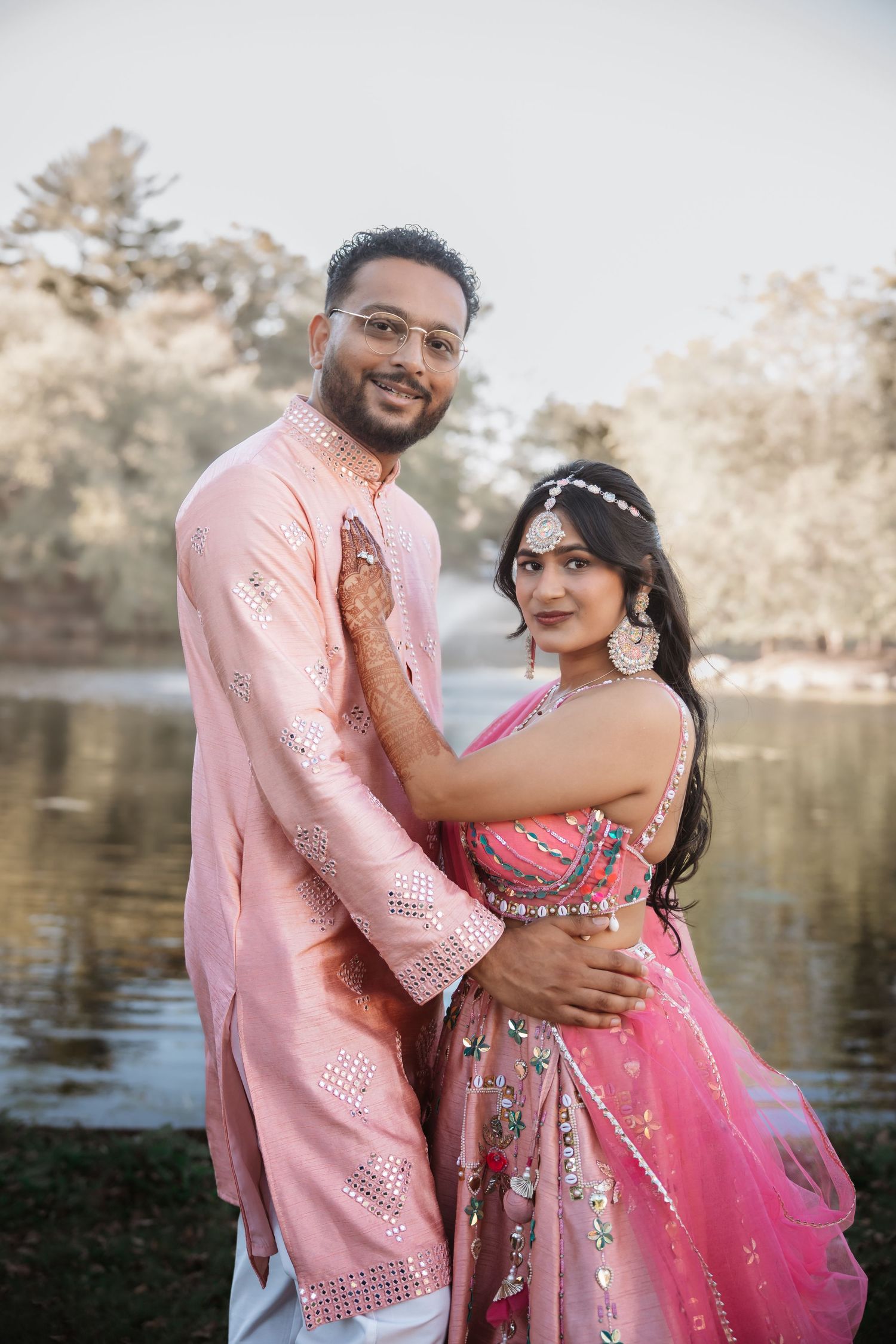 Indian couple in matching pink traditional wedding outfits pose lovingly together near a serene lake backdrop.