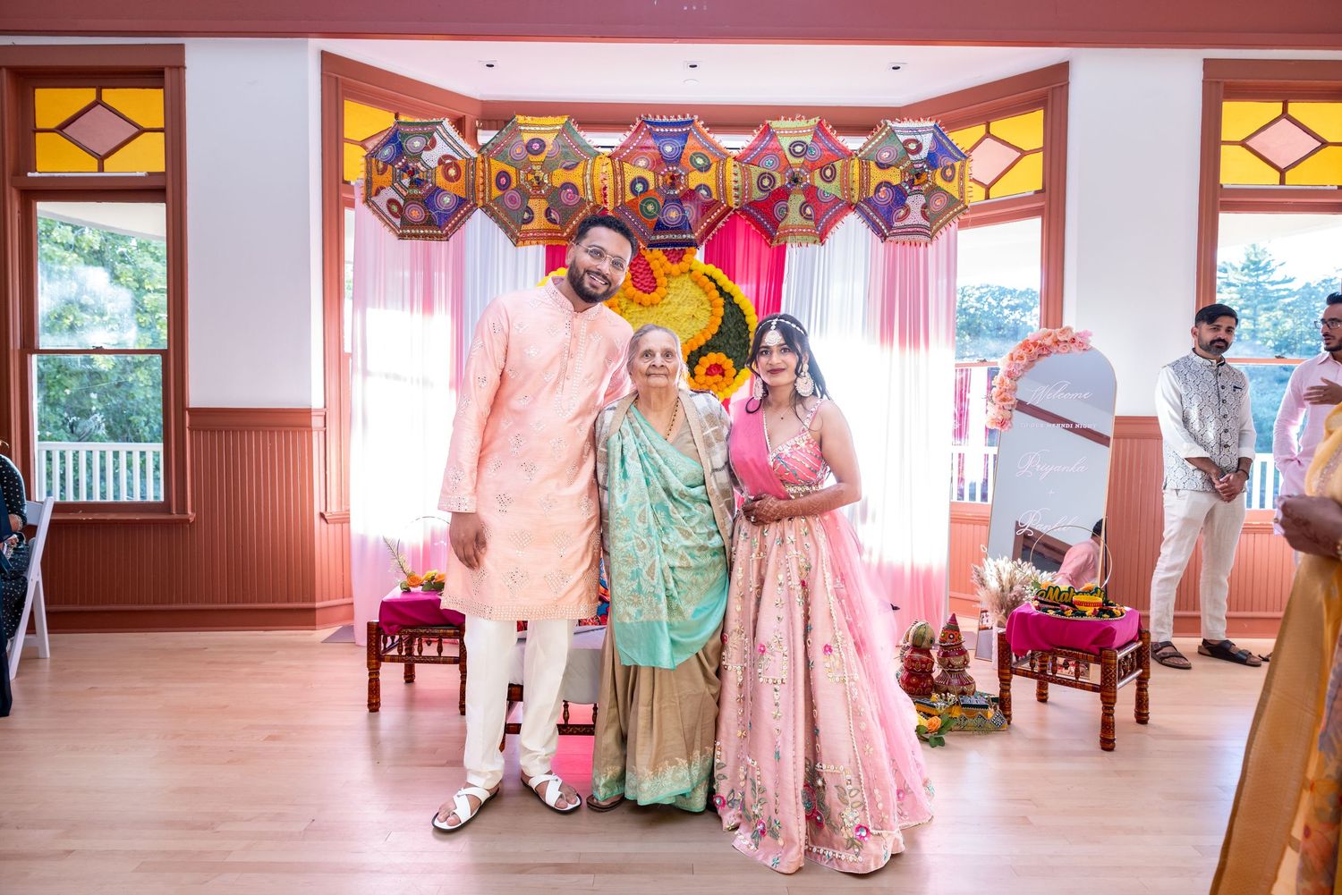 Indian bride in pink lehenga poses with groom and elder at decorated wedding venue.