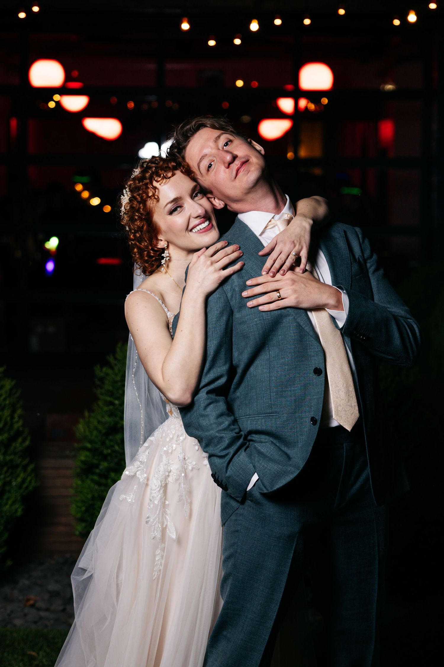 Bride in white lace gown hugs groom in gray suit outdoors at night, smiling warmly under string lights.