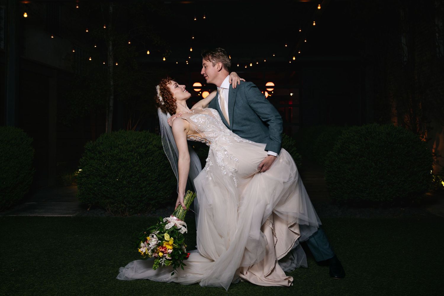 Groom dips bride in lace gown for romantic nighttime wedding photo surrounded by string lights.