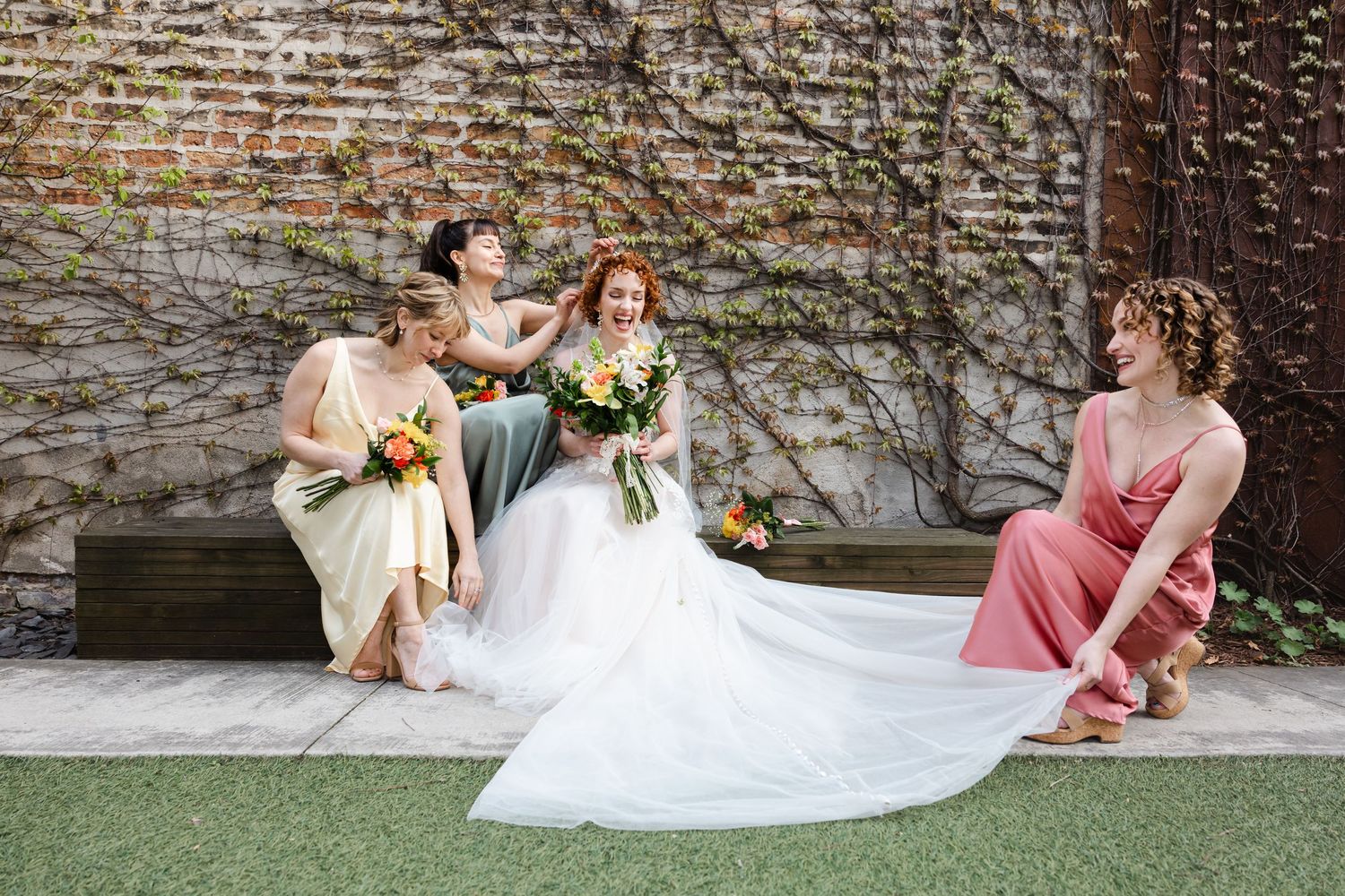 Bride in white gown sits with three bridesmaids in colorful dresses against ivy-covered stone wall outdoors.