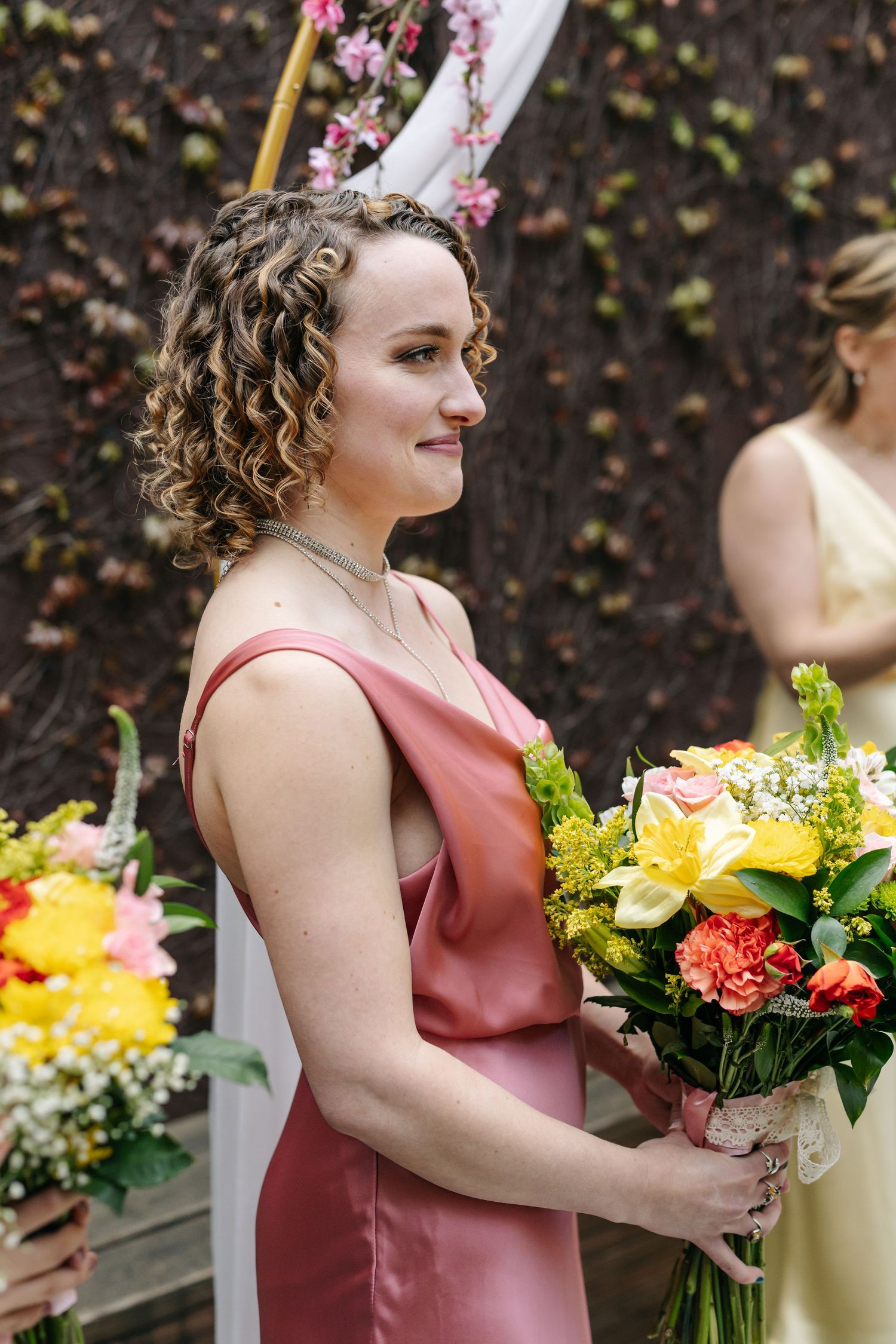 Bridesmaid in pink satin dress holding colorful bouquet of yellow and orange flowers at wedding ceremony.
