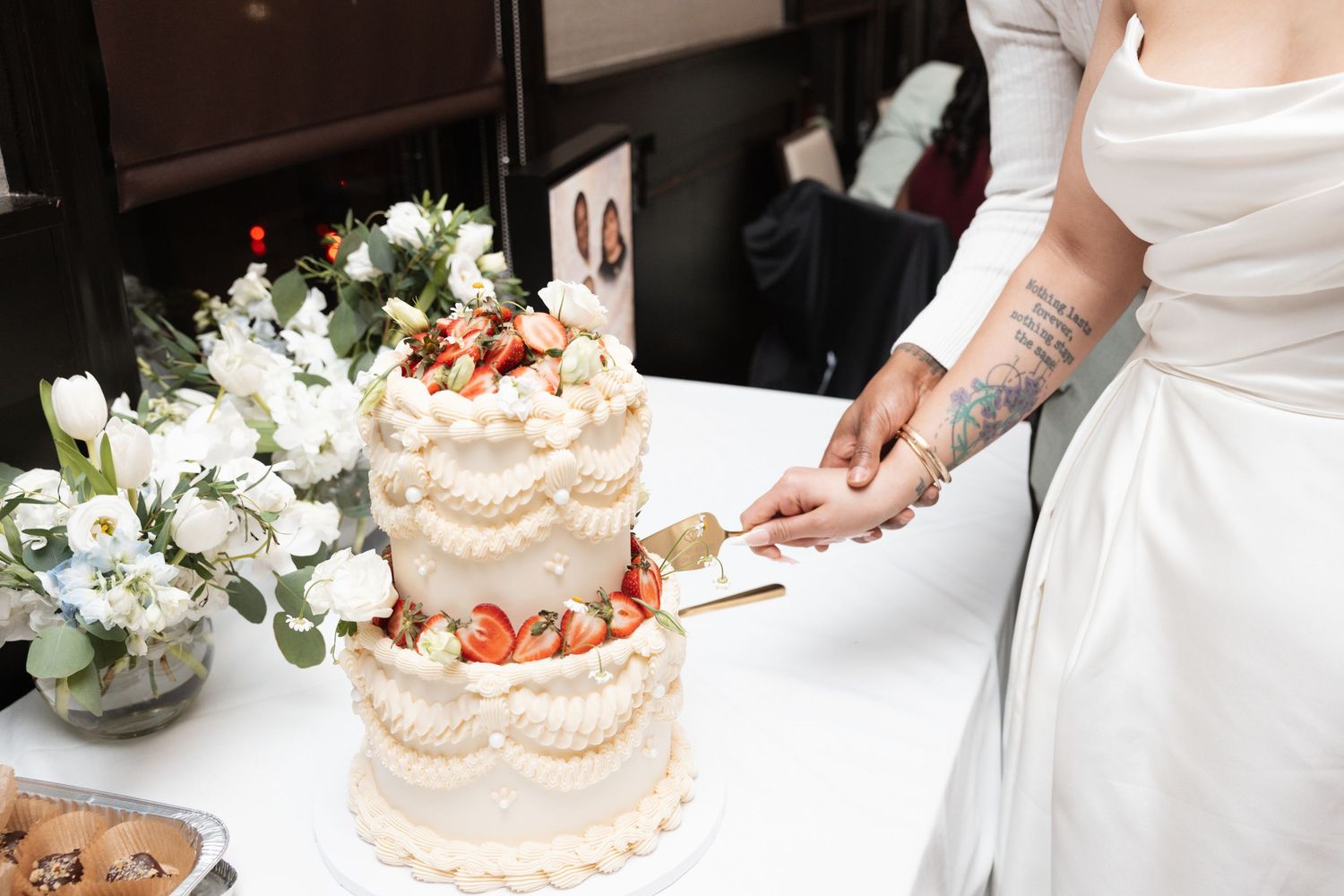 Bride with tattoos cutting a two-tier white wedding cake decorated with fresh strawberries.