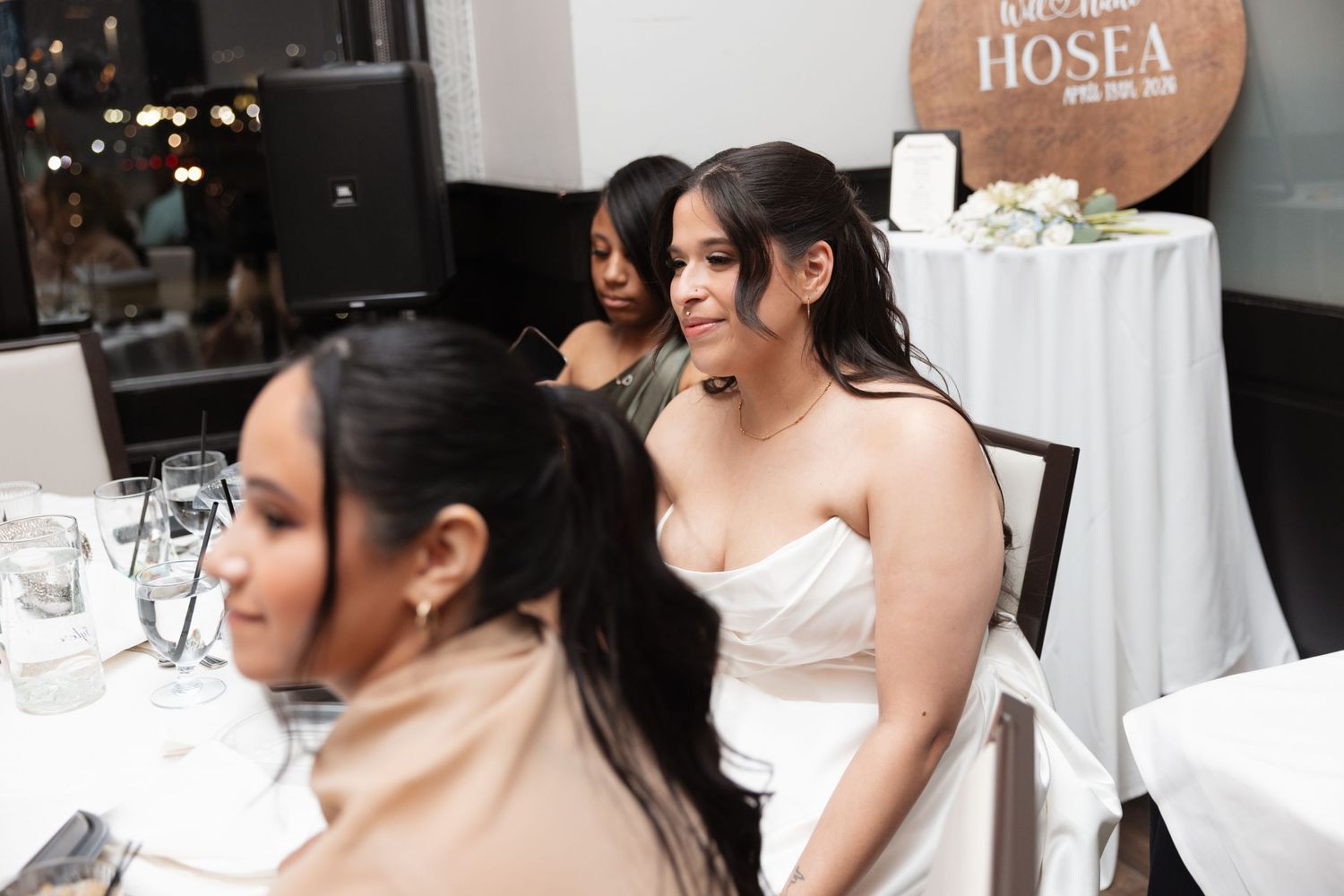 Bride in white strapless dress sits at decorated wedding reception table smiling at guests.