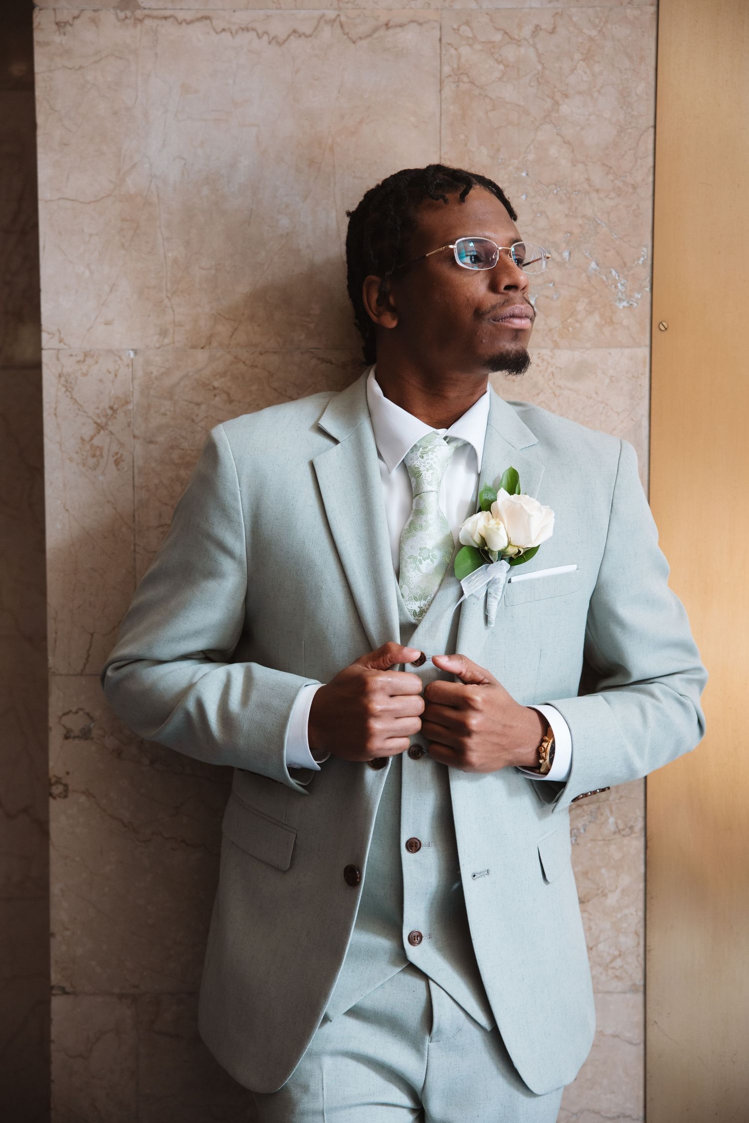 Elegant groom in light gray three-piece suit with boutonniere leans against marble wall at wedding.