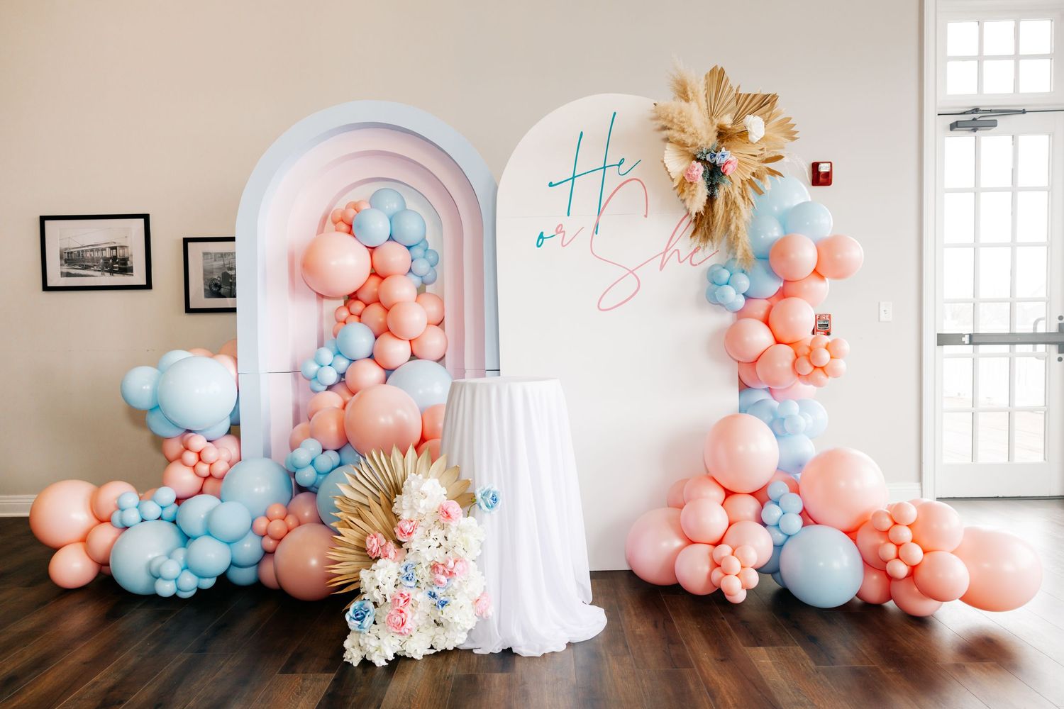 Gender reveal party backdrop with pink and blue balloon garland arch display and He or She sign.