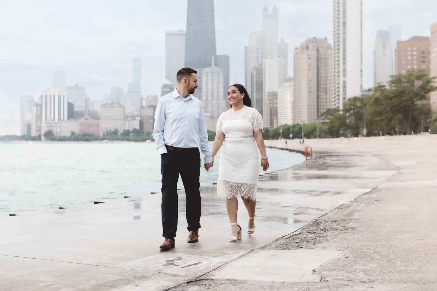 Couple holding hands walking along Chicago lakefront with city skyline in background on cloudy day.