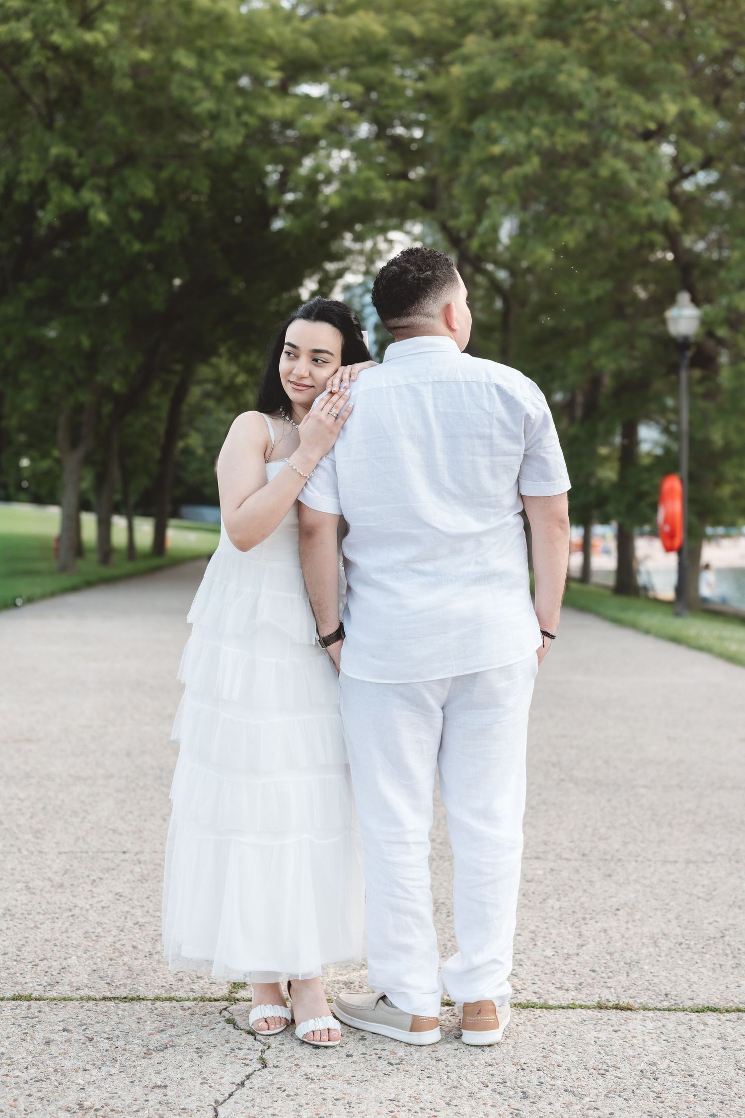 Woman in white tiered dress holding mans arm as they walk together along a tree-lined path.