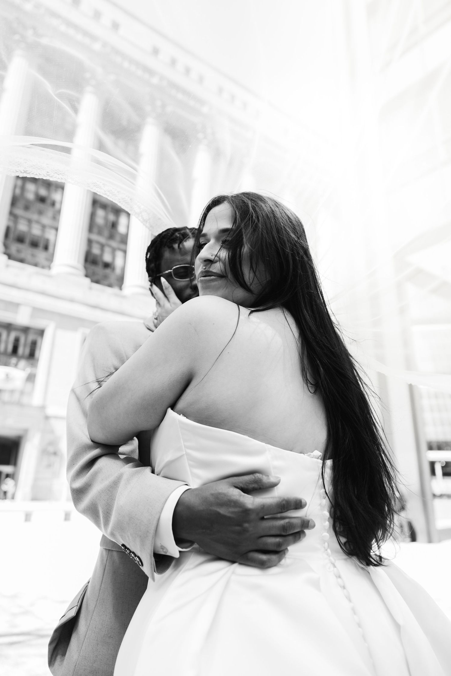 Black and white photo of bride and groom sharing tender embrace outside grand building.
