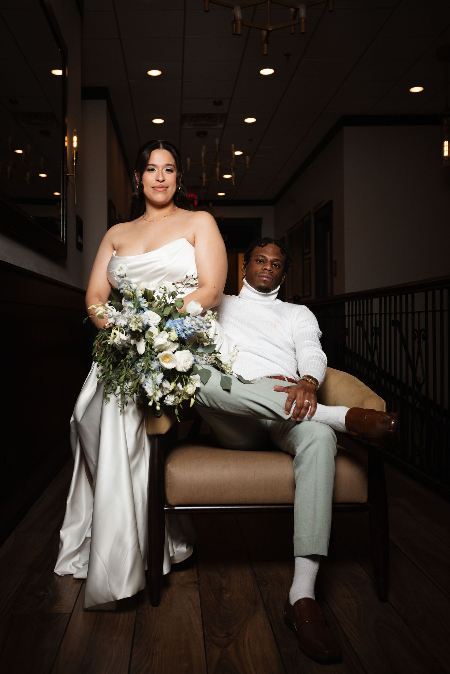 Bride standing beside groom seated in chair, holding blue and white floral bouquet in elegant dark venue.