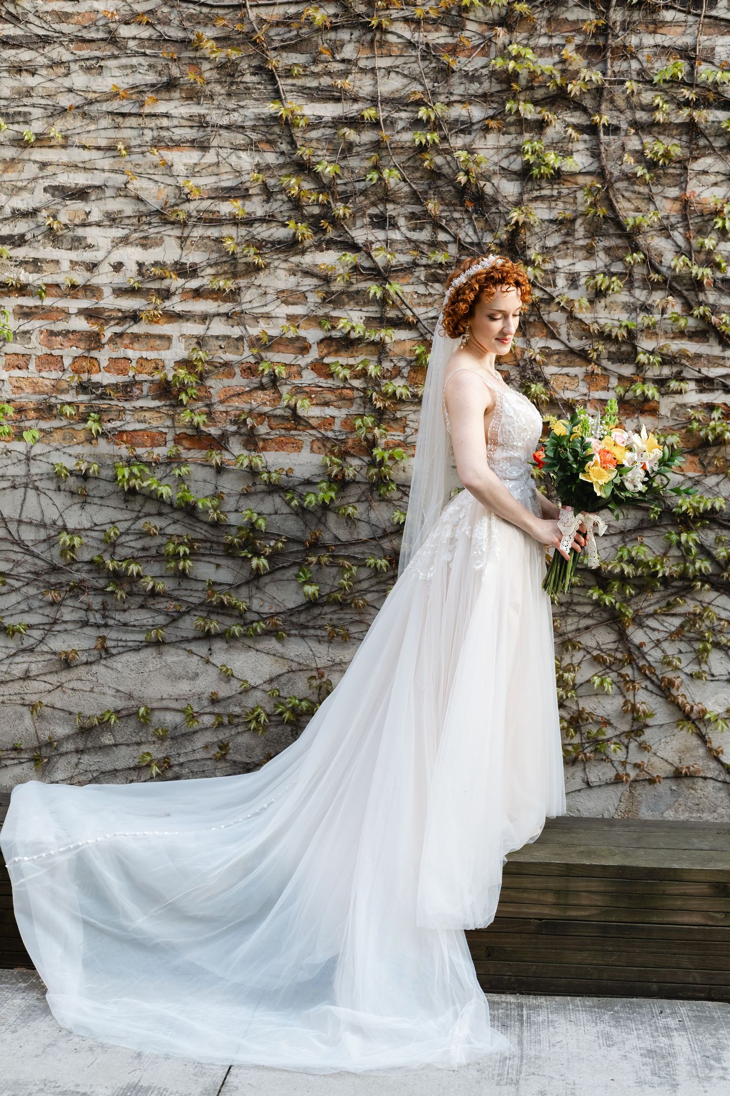 Elegant bride in white lace gown with floral crown holding colorful bouquet in front of ivy-covered brick wall.