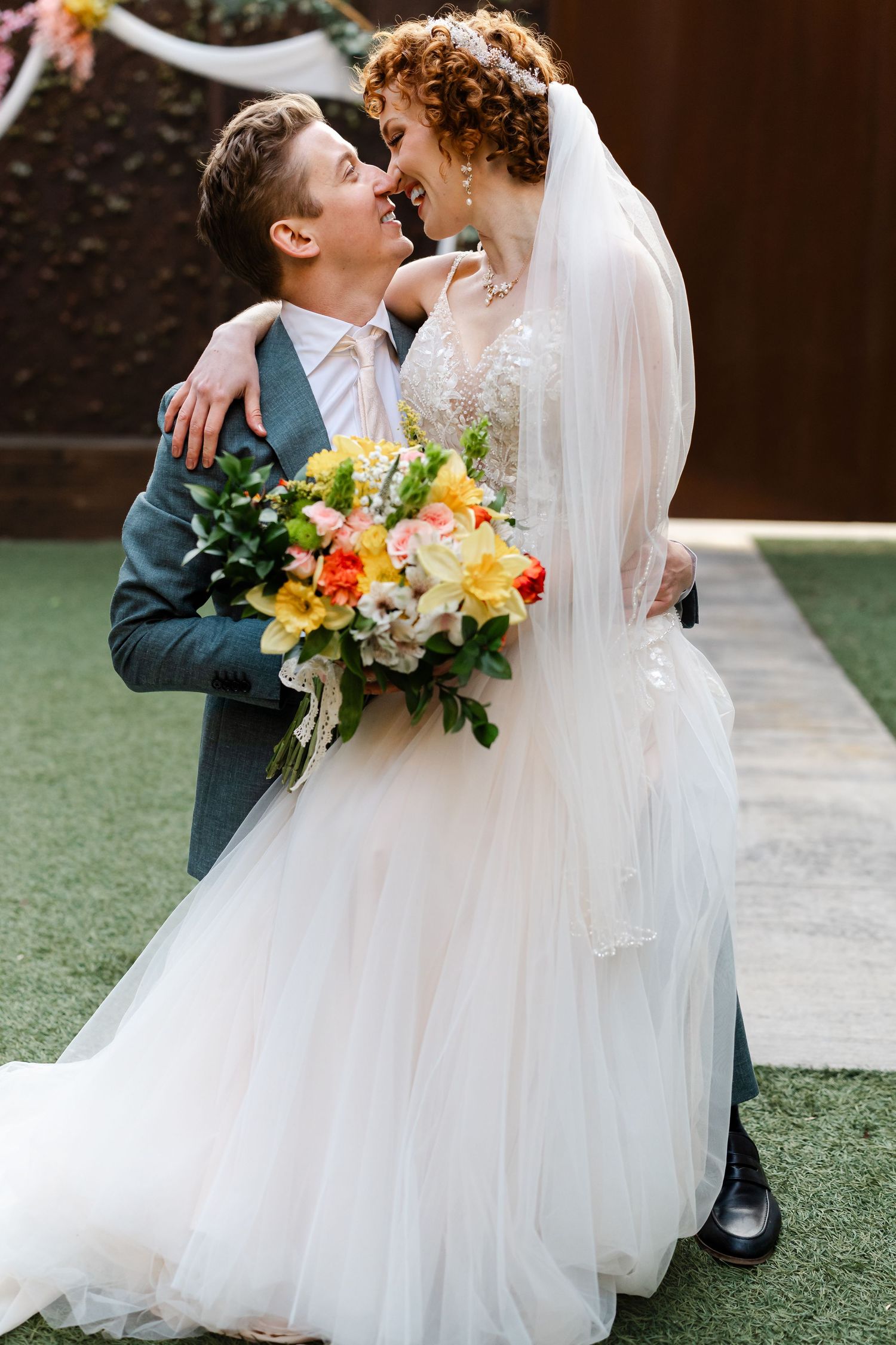 Joyful bride and groom in a tender moment, she in a white tulle gown holding a vibrant yellow and orange bouquet.