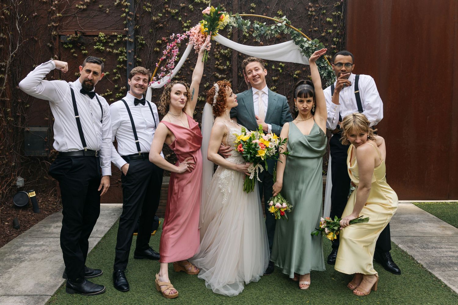 Wedding party celebrates under floral arch with bridesmaids in colorful dresses and groomsmen.
