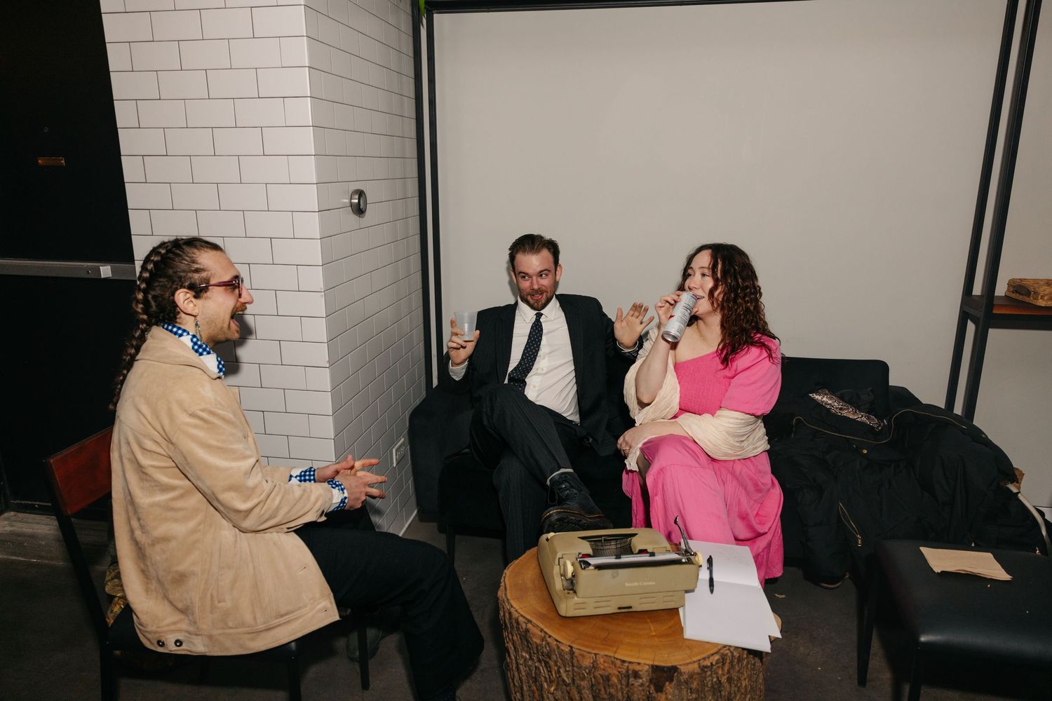 Three wedding guests laugh and chat in cozy lounge area with vintage typewriter centerpiece at reception.