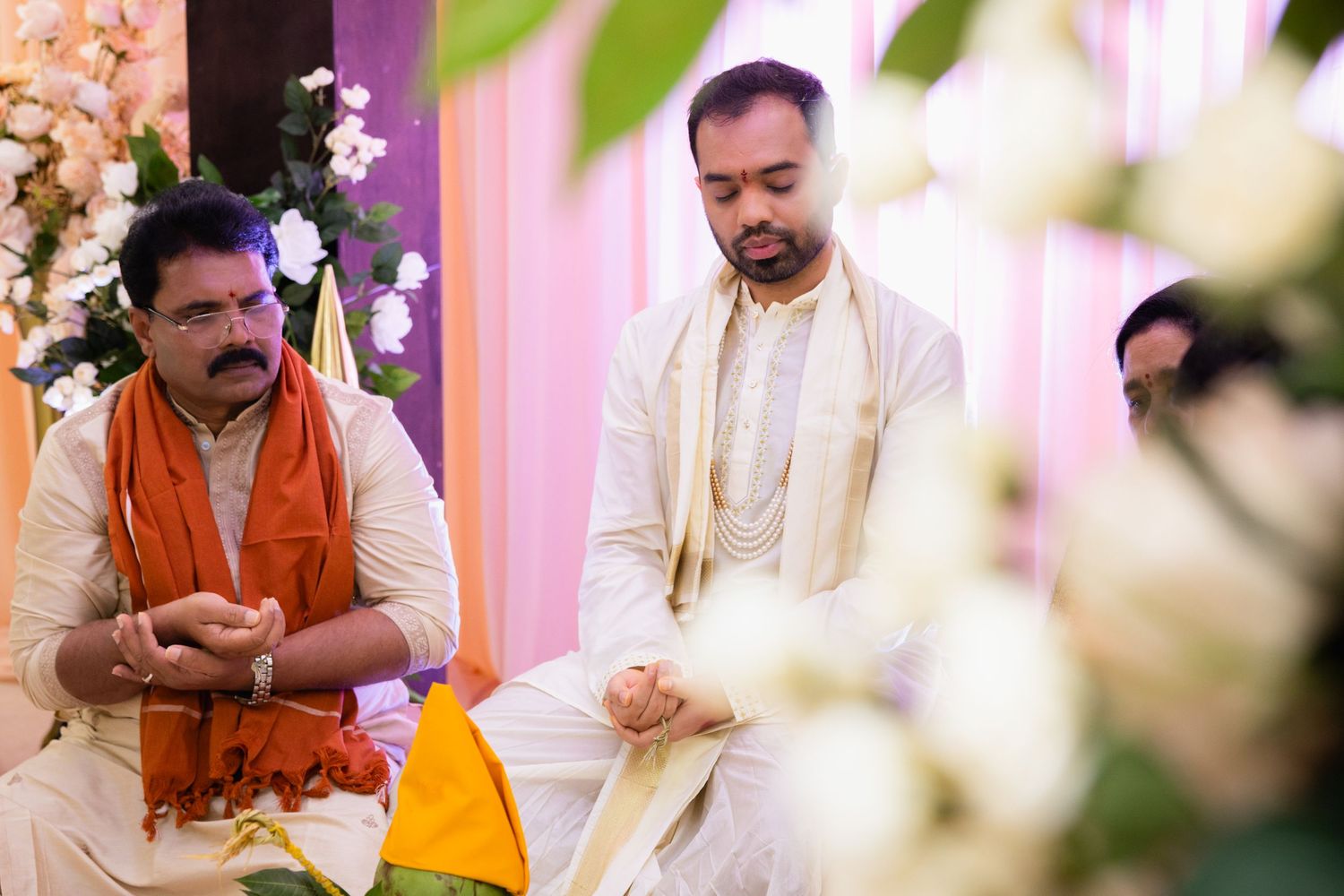 Indian groom in white kurta seated during Hindu wedding ceremony with priest in orange attire.