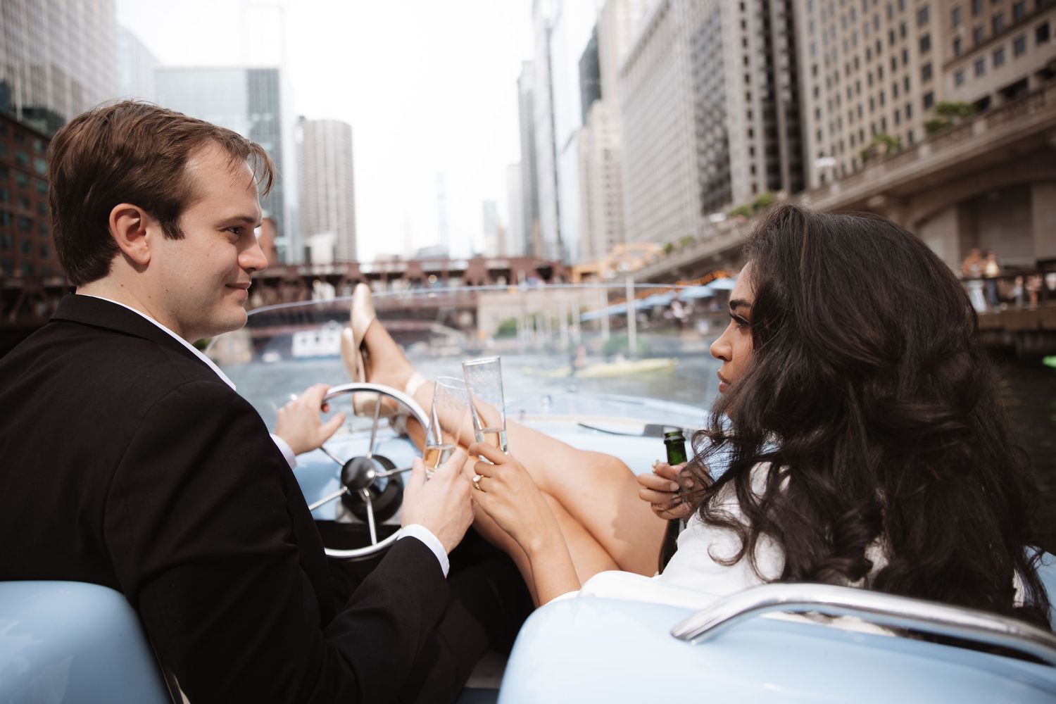 Couple in formal attire sharing a romantic moment in a convertible car along a Chicago river cityscape.