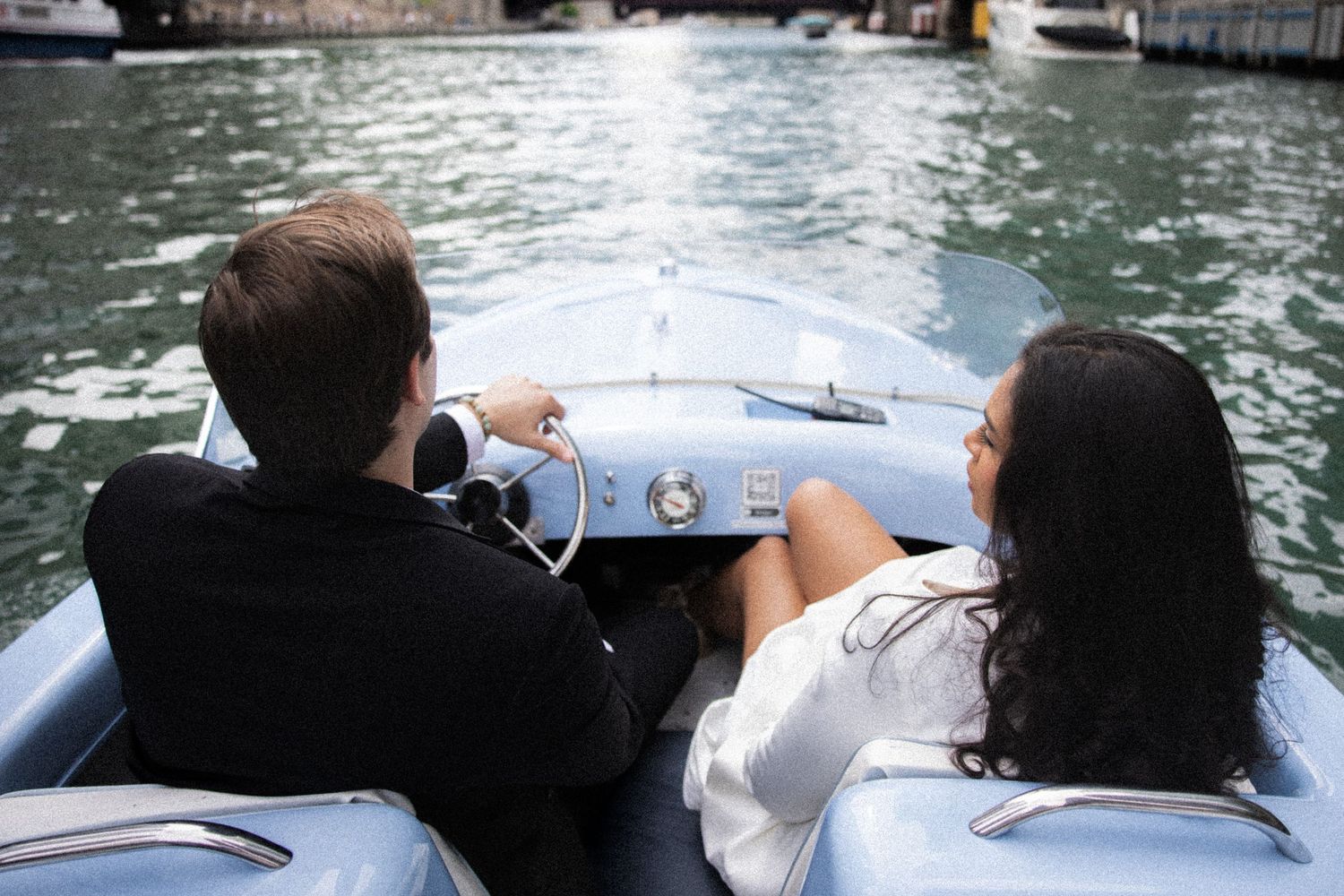 Couple riding in a vintage light blue speedboat on calm water, viewed from behind.