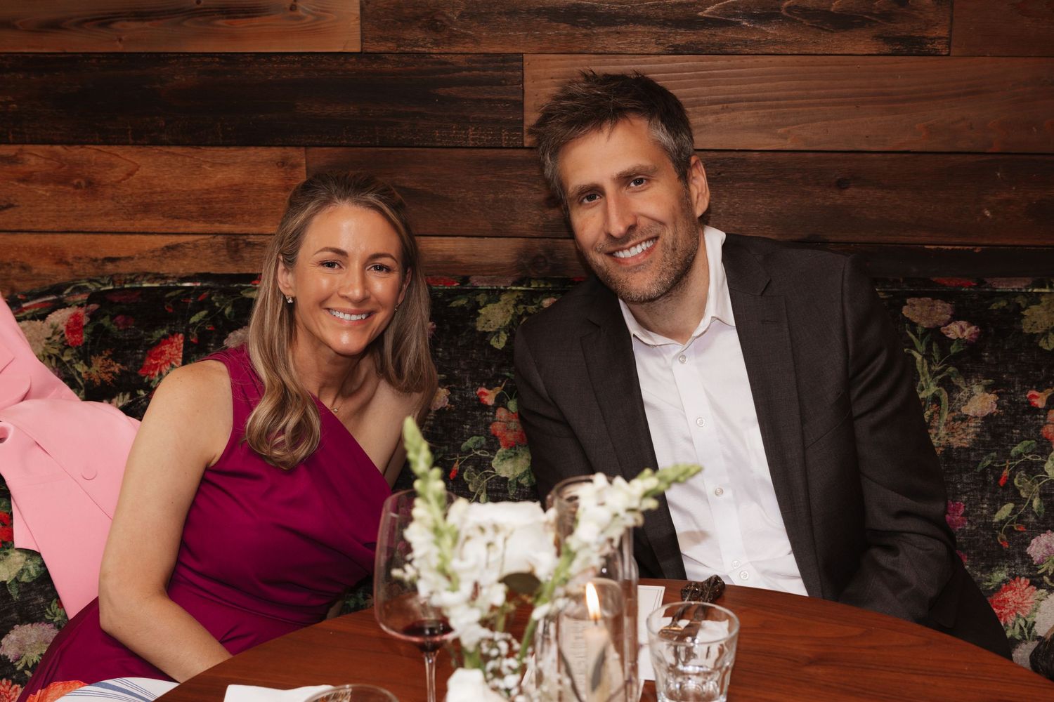 Smiling couple poses together on a floral booth at a cozy wedding reception dinner table.