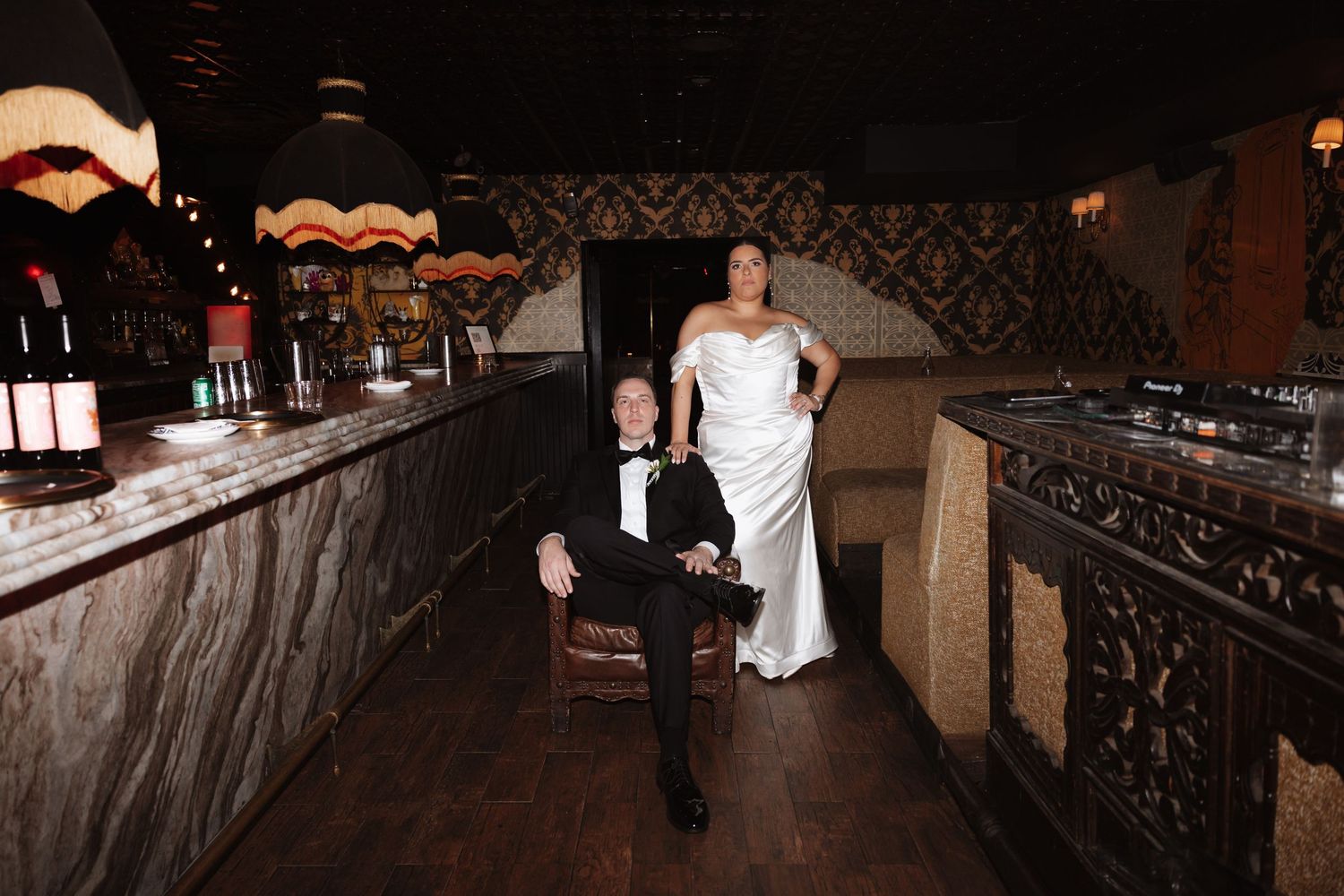 Bride in white gown and groom in tuxedo pose in elegant dark bar venue on their wedding day.