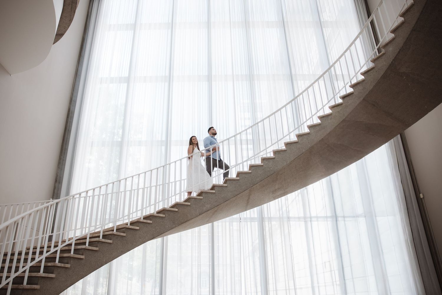 Couple in wedding attire standing on a curved white staircase in a modern building with floor-to-ceiling windows.