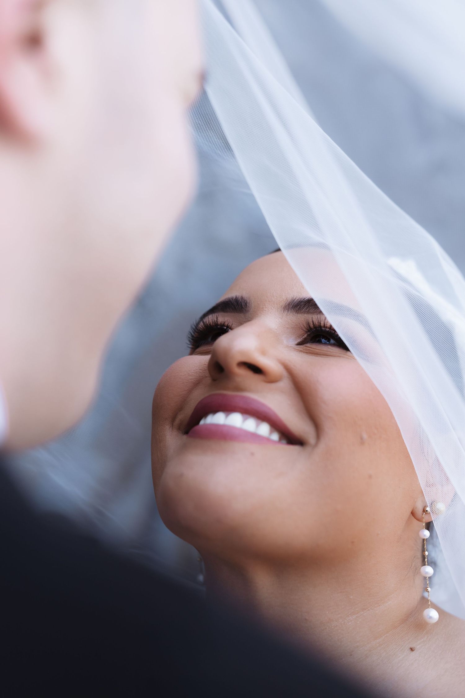 Smiling bride gazes up at groom under flowing white veil, wearing pearl drop earrings.