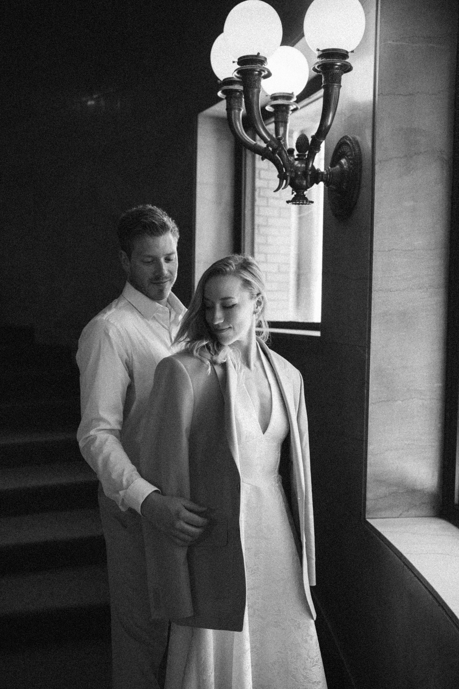 Black and white photo of groom placing jacket on bride near ornate wall sconce.