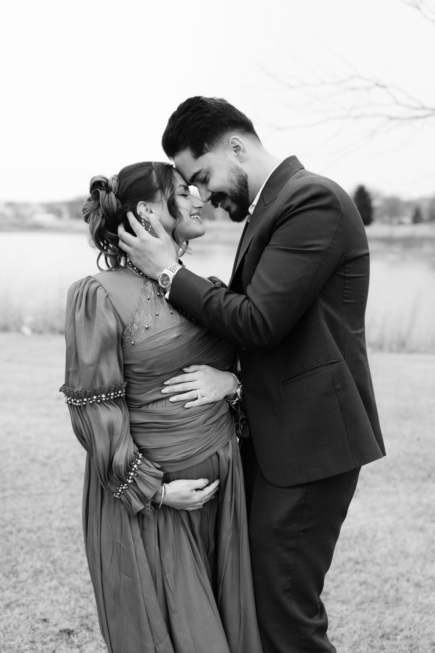 Black and white romantic maternity photo of couple touching foreheads near a peaceful lake.
