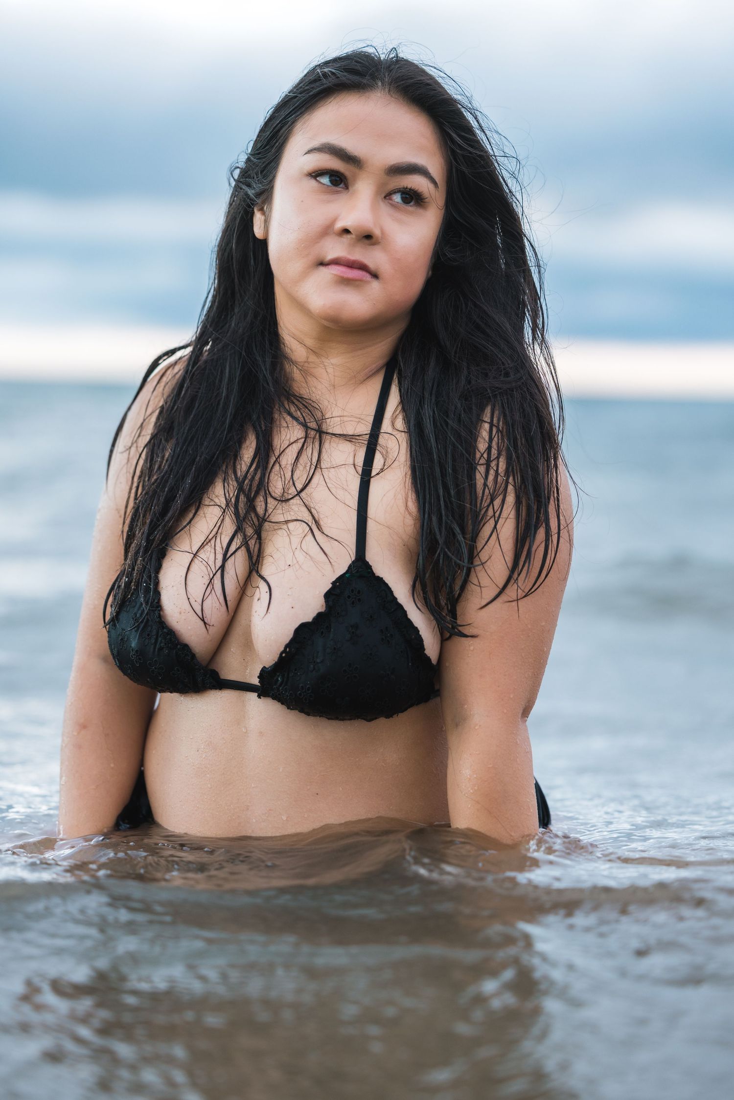 Woman in black bikini sitting in shallow ocean water with long dark wet hair and cloudy sky.
