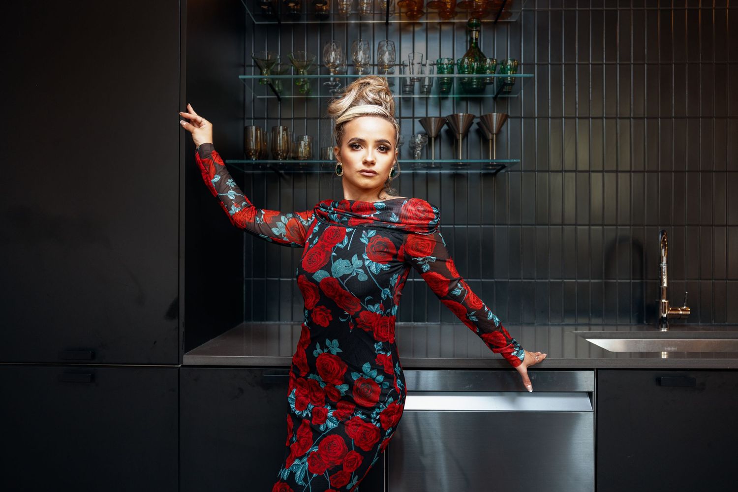 Woman wearing floral red rose gown with crown accessory posing in modern dark kitchen.