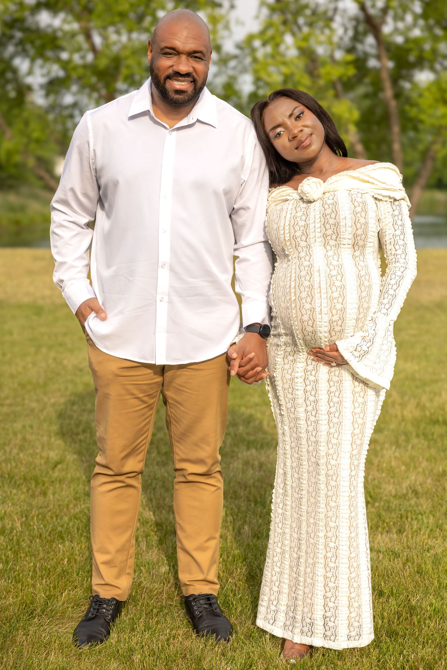 A smiling pregnant couple poses together on a green lawn.