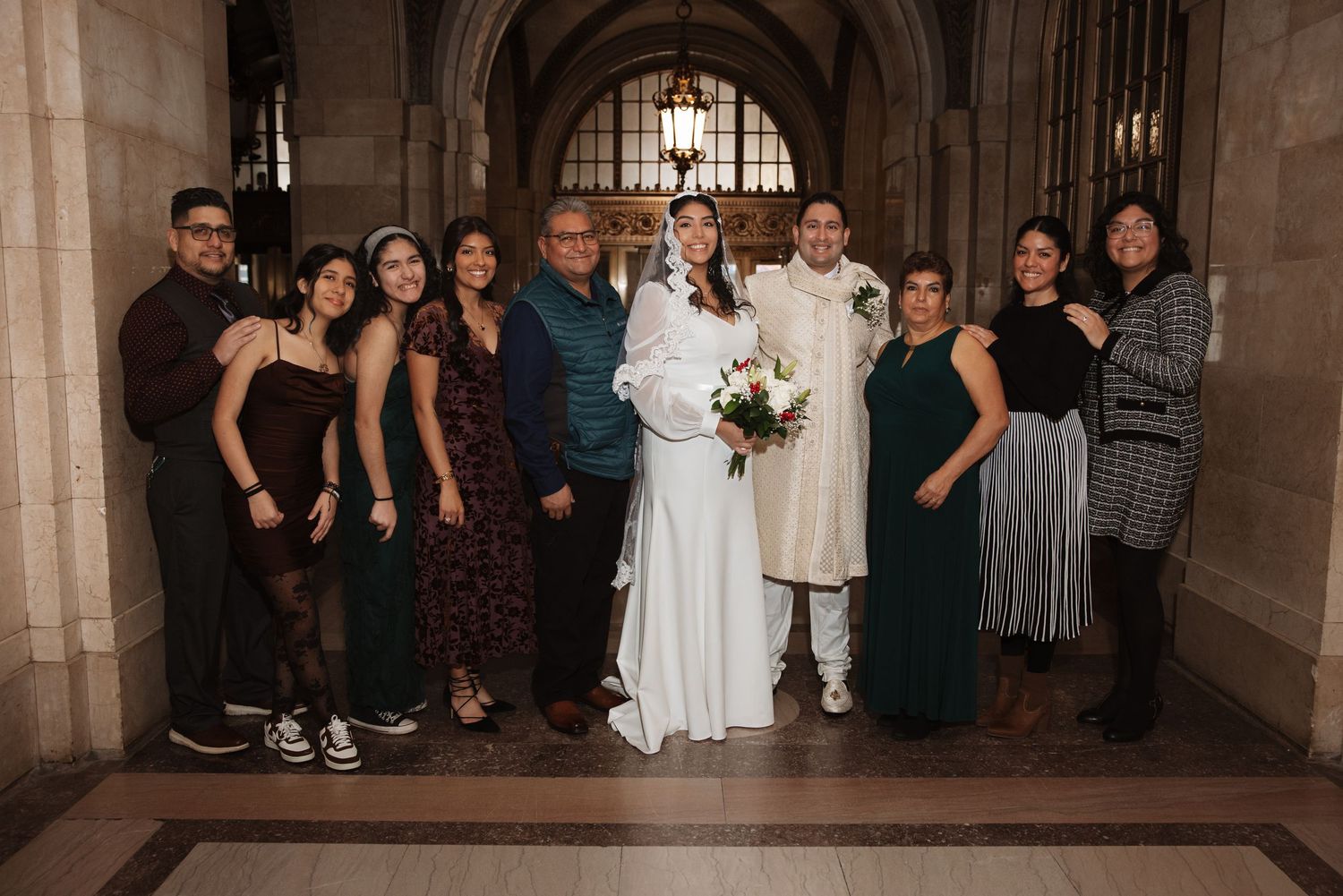 Bride in white gown and groom in ivory sherwani pose with large family group in ornate cathedral hallway.