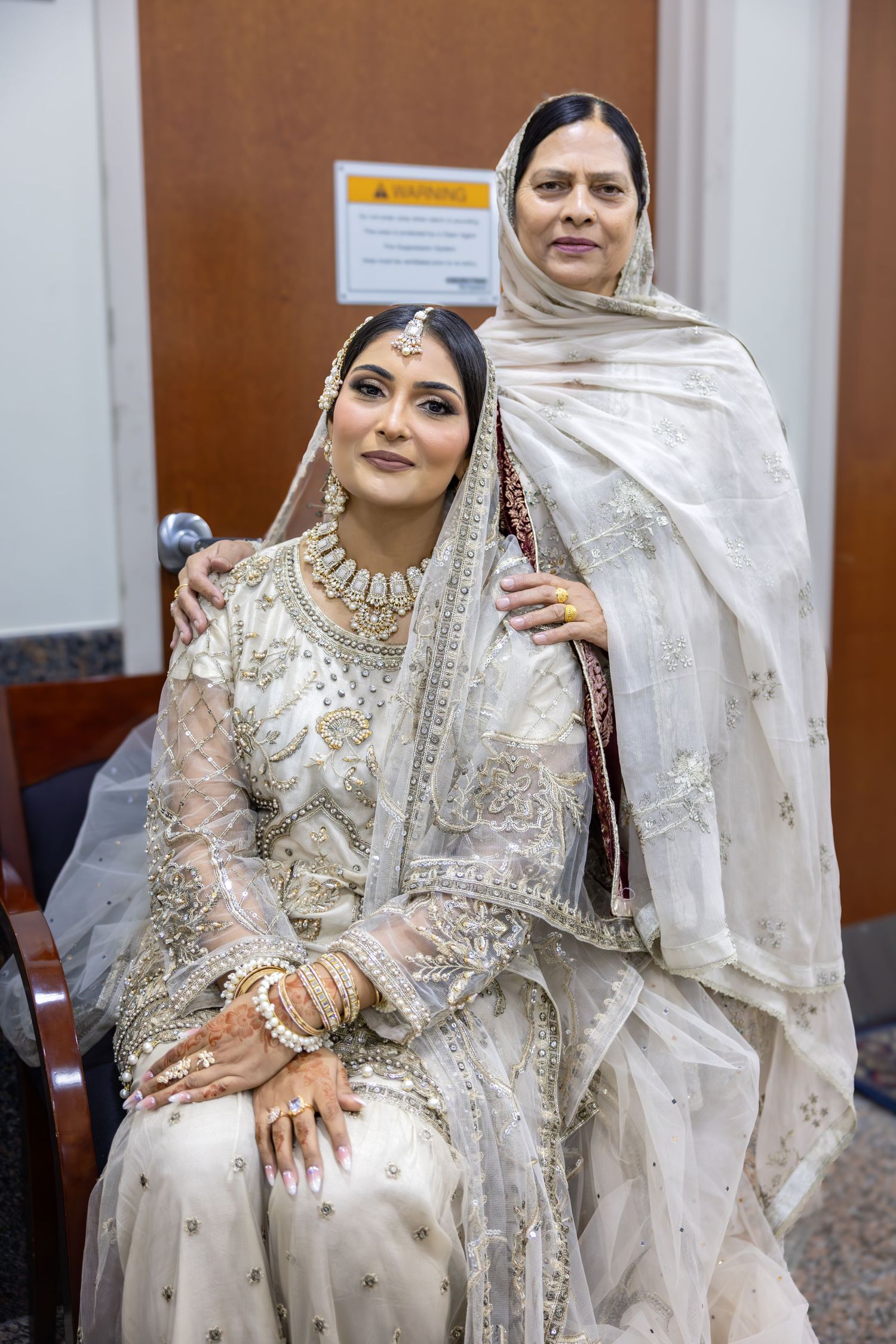 A bride in white embroidered South Asian bridal attire sits with an older woman in a hijab standing behind her.