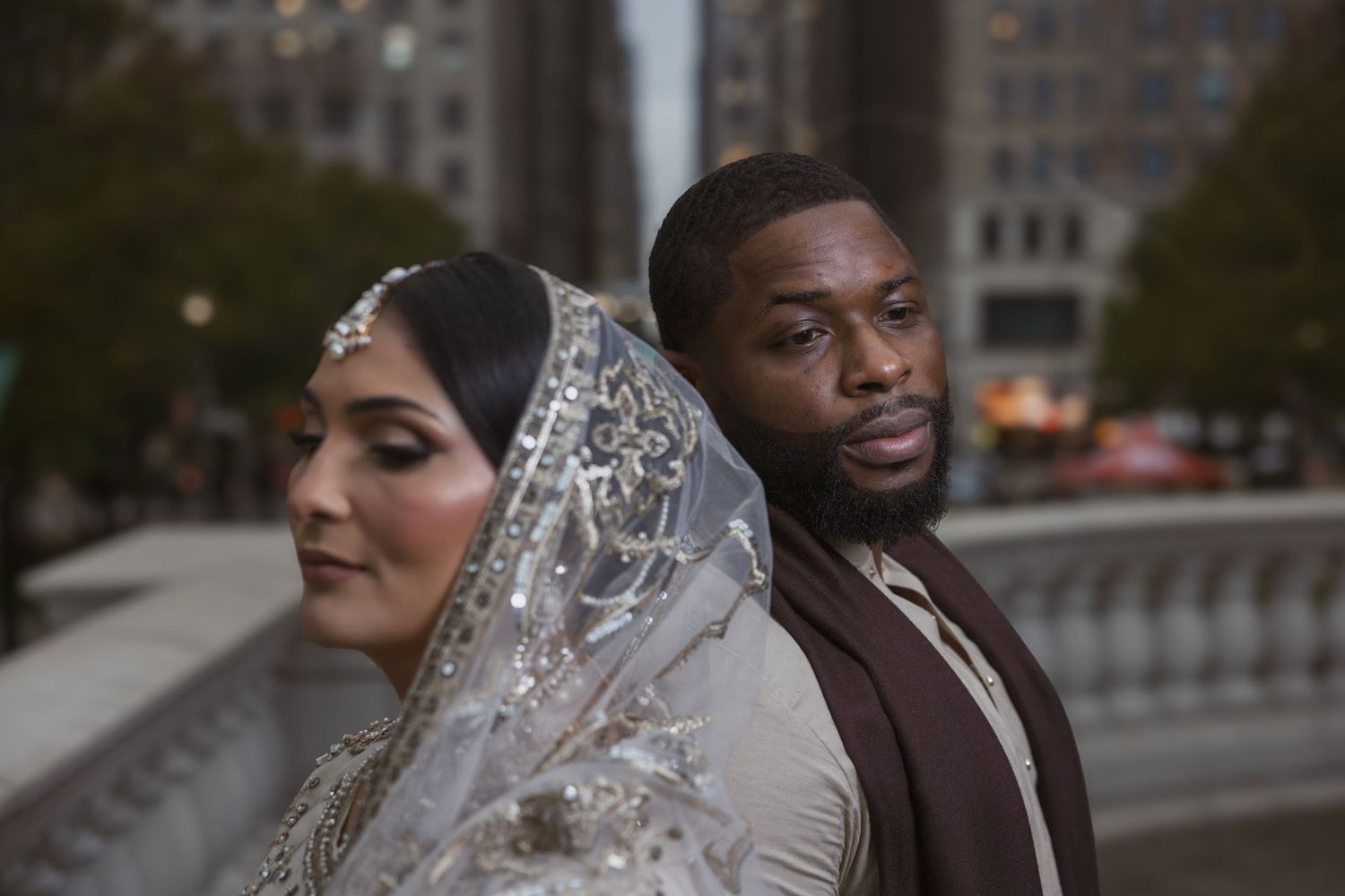 Bride in silver embroidered dupatta and groom in brown sherwani posing back-to-back outdoors.