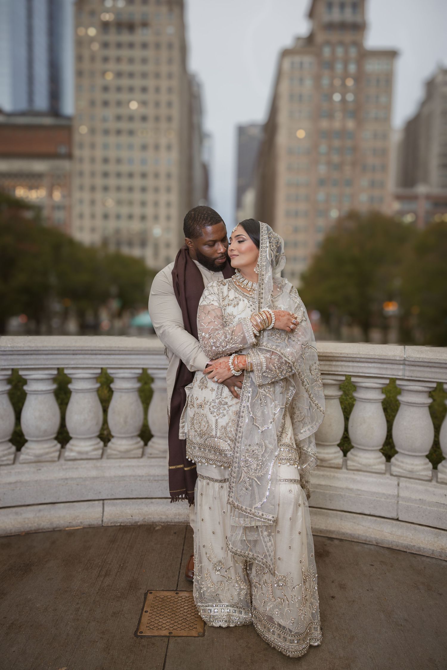 Groom in beige sherwani tenderly embraces bride in ivory lehenga on a stone balcony with city backdrop.