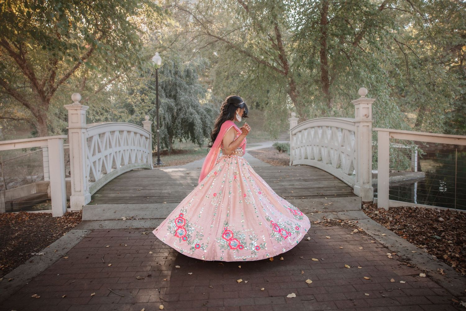 Bride showcasing floral pink lehenga from behind on scenic white arched garden bridge.