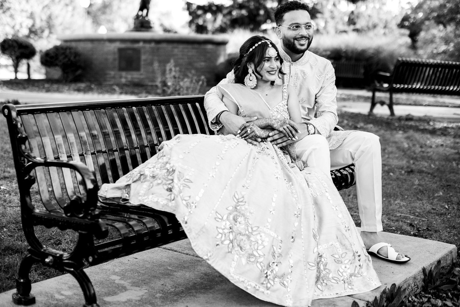 Black and white photo of Indian couple sitting on park bench in wedding attire.