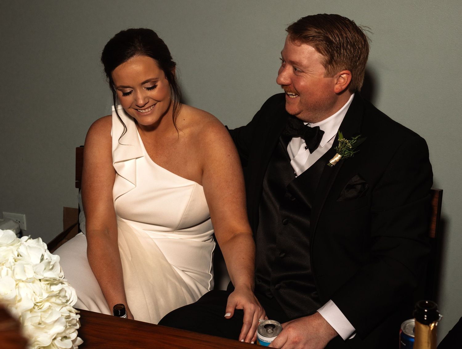 Bride in white one-shoulder dress and groom in tuxedo laugh together while signing wedding documents.