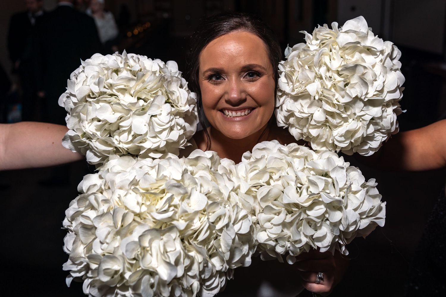 Joyful bridesmaid playfully hiding behind four large white hydrangea bouquets at wedding reception.