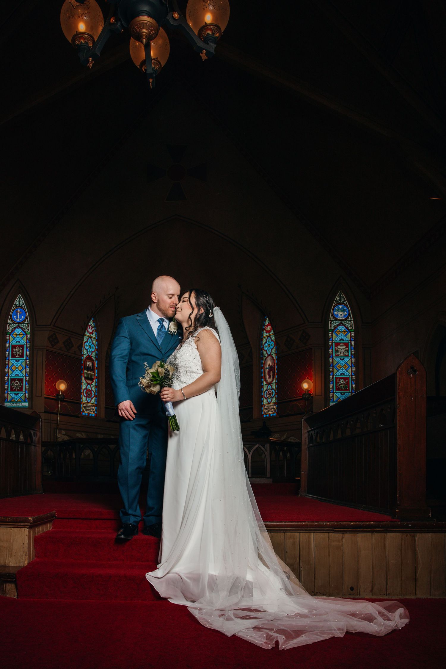 Newlyweds share a romantic kiss at the altar of a dimly lit church with colorful stained glass windows.