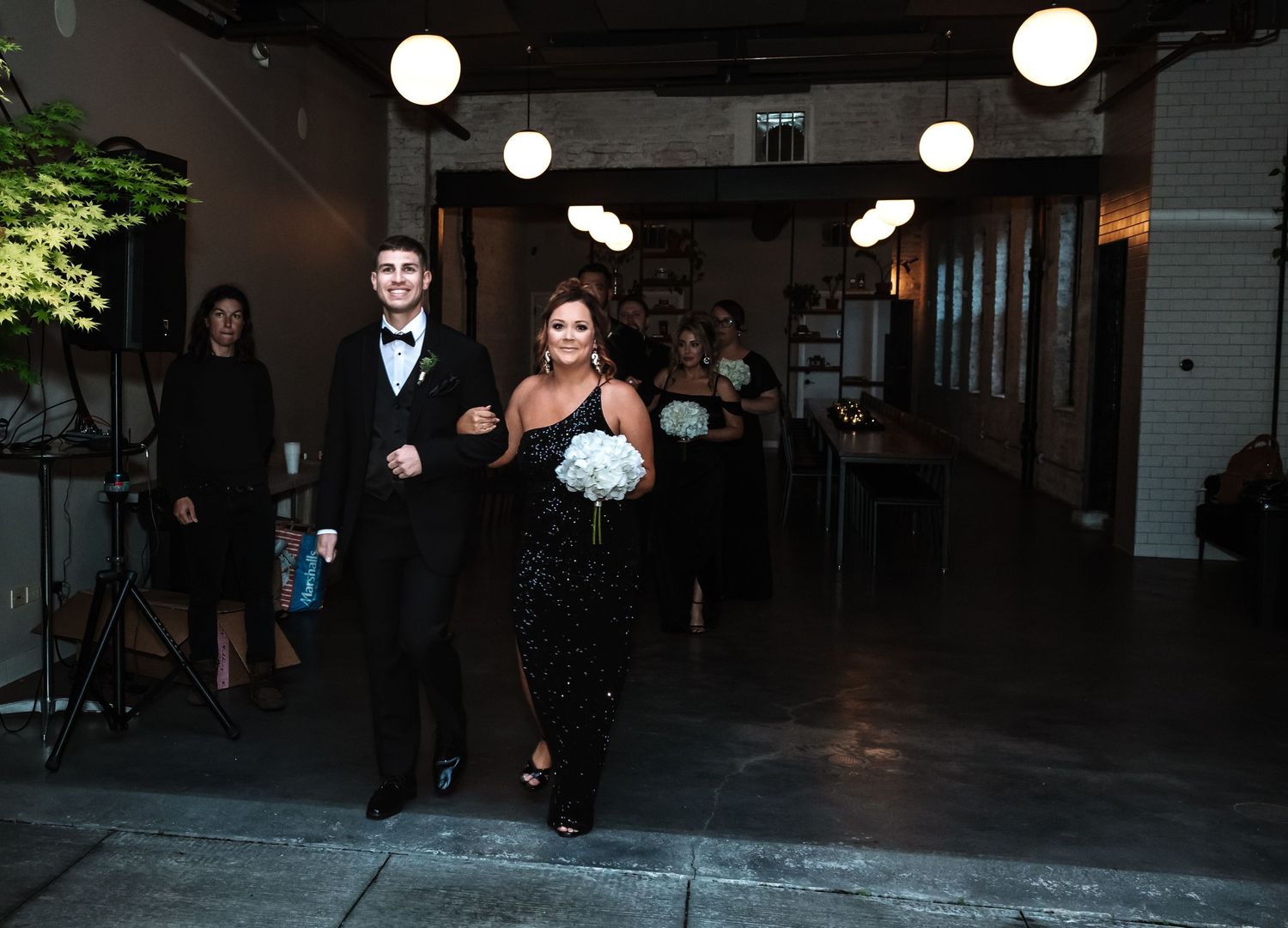 Groomsman in tuxedo escorting bridesmaid in black sequin dress holding white bouquet.
