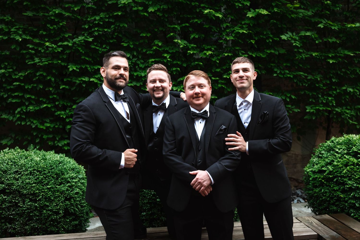 Four men in black tuxedos smiling together outdoors near trimmed hedges and a green ivy-covered wall.