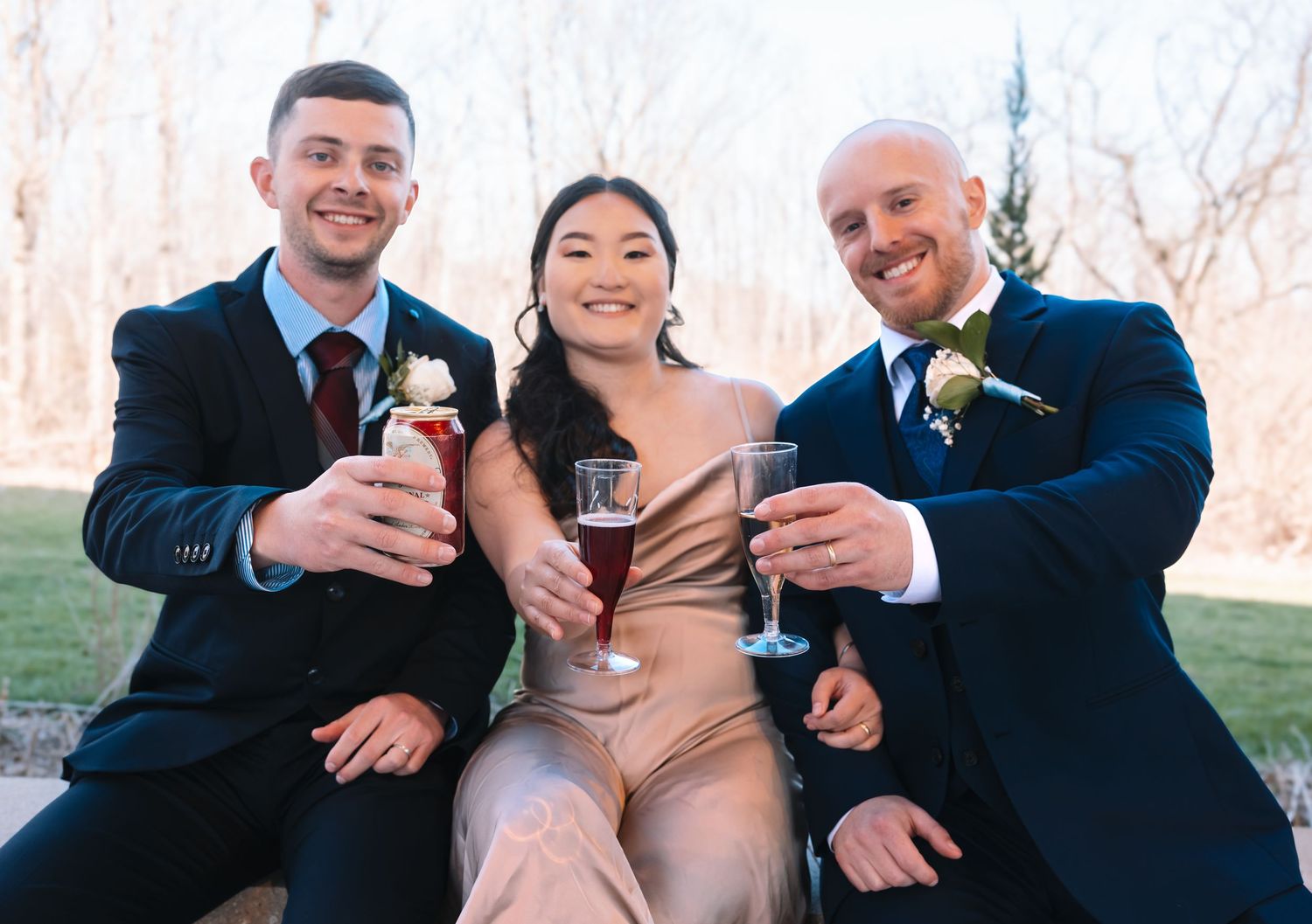 Three wedding guests in formal attire smiling and raising champagne glasses during an outdoor celebration.