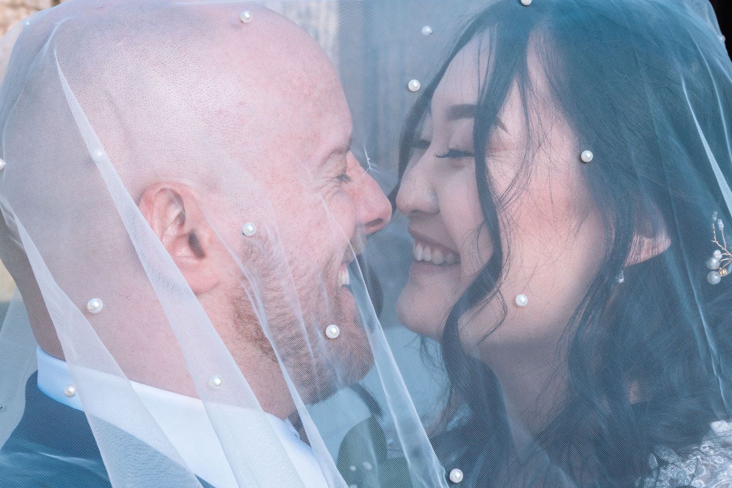 Laughing bride and bald groom share a joyful nose-to-nose moment under a pearl-dotted wedding veil.