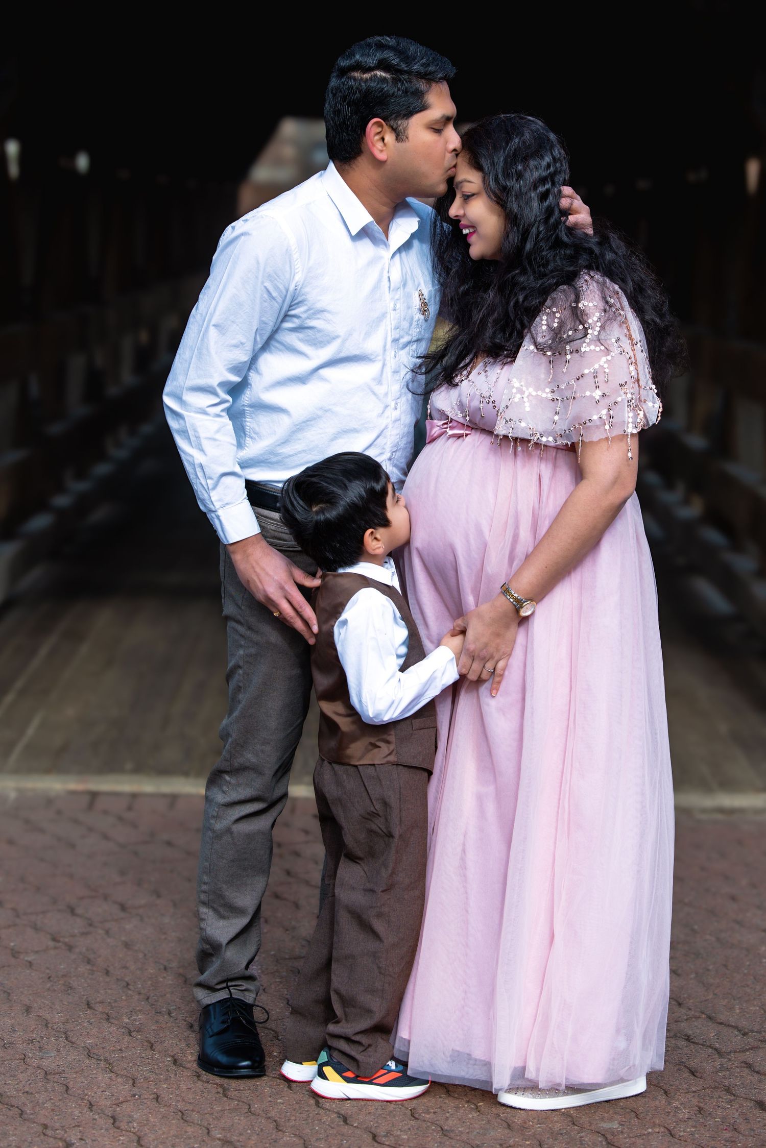 Tender family maternity photo of Indian couple with young son kissing mothers belly under a covered wooden bridge.