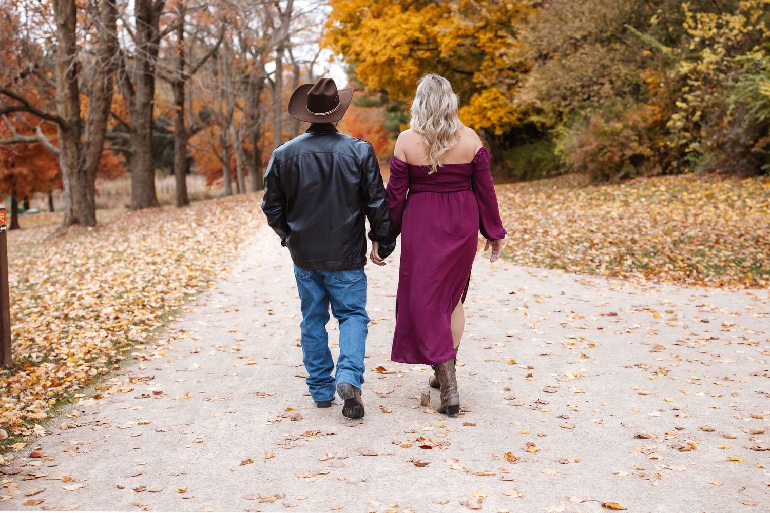 Pregnant woman in burgundy dress and cowboy partner walking away on autumn park path.
