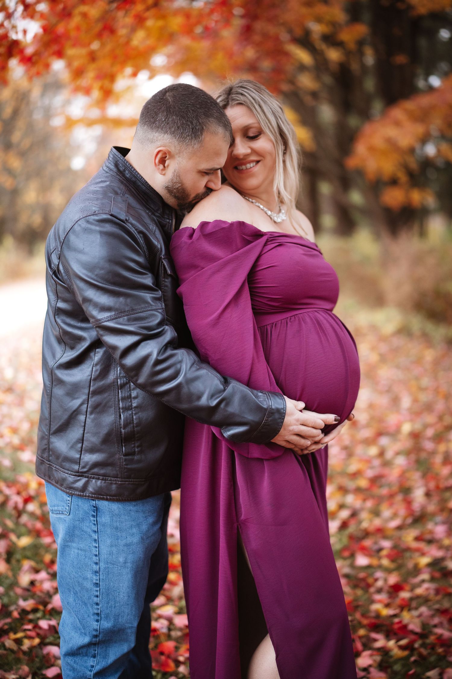 Man in leather jacket embracing smiling pregnant woman in purple maternity dress in fall setting.