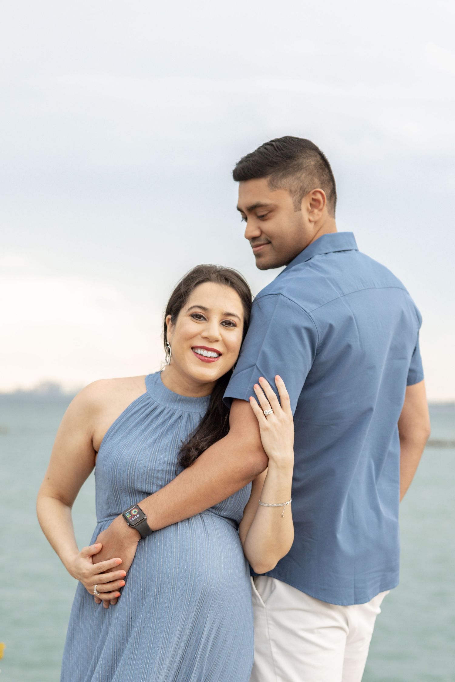 Happy pregnant woman in blue dress embraced by partner near ocean backdrop.