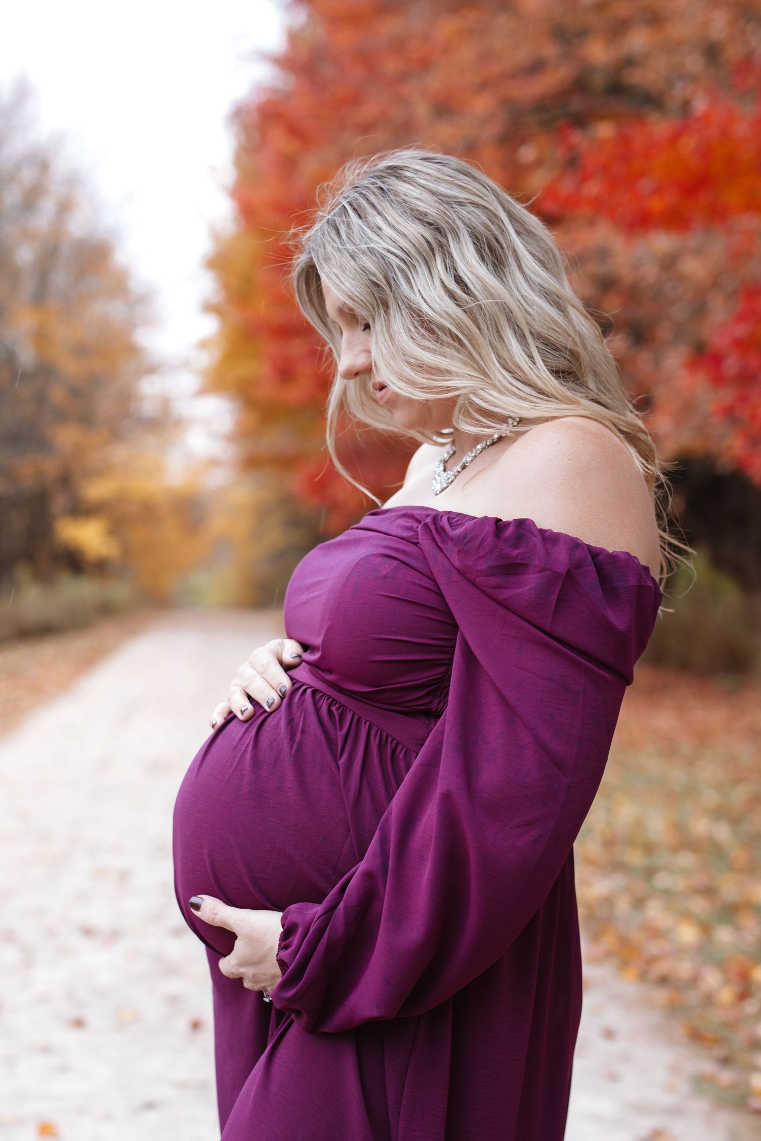 Blonde pregnant woman in purple maternity dress gently cradling bump on autumn tree-lined path.